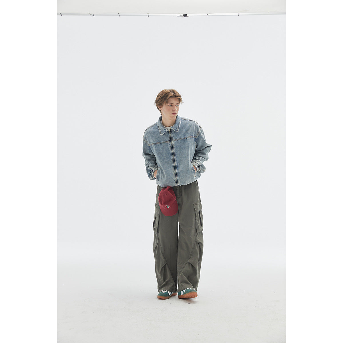 Model wearing a light-wash blue denim zip jacket and loose dark cargo pants, with a red cap at his side.