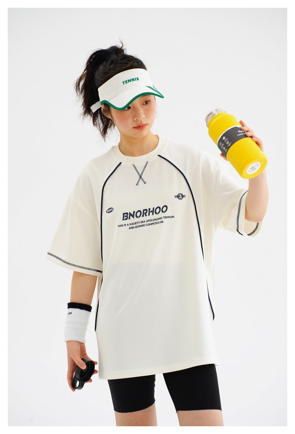 Close view of a model wearing an ivory oversized T-shirt with dark piping and printed chest graphics, with black shorts.