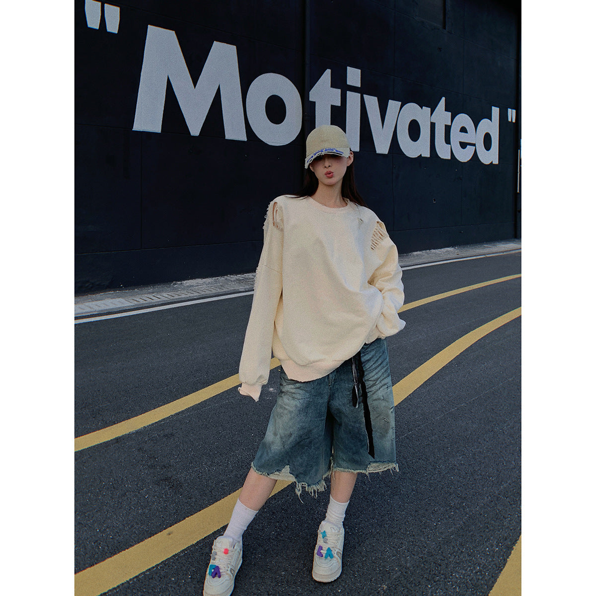 Model wearing an oversized cream distressed sweatshirt and frayed denim shorts with a cap and sneakers on a street.