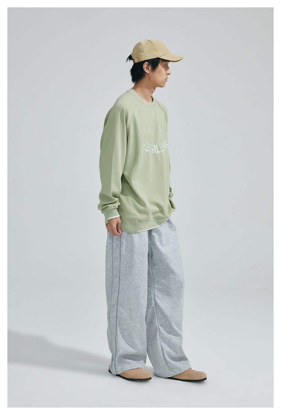 Model in side view wearing a light green oversized crew-neck sweatshirt with gray wide-leg sweatpants and a tan cap