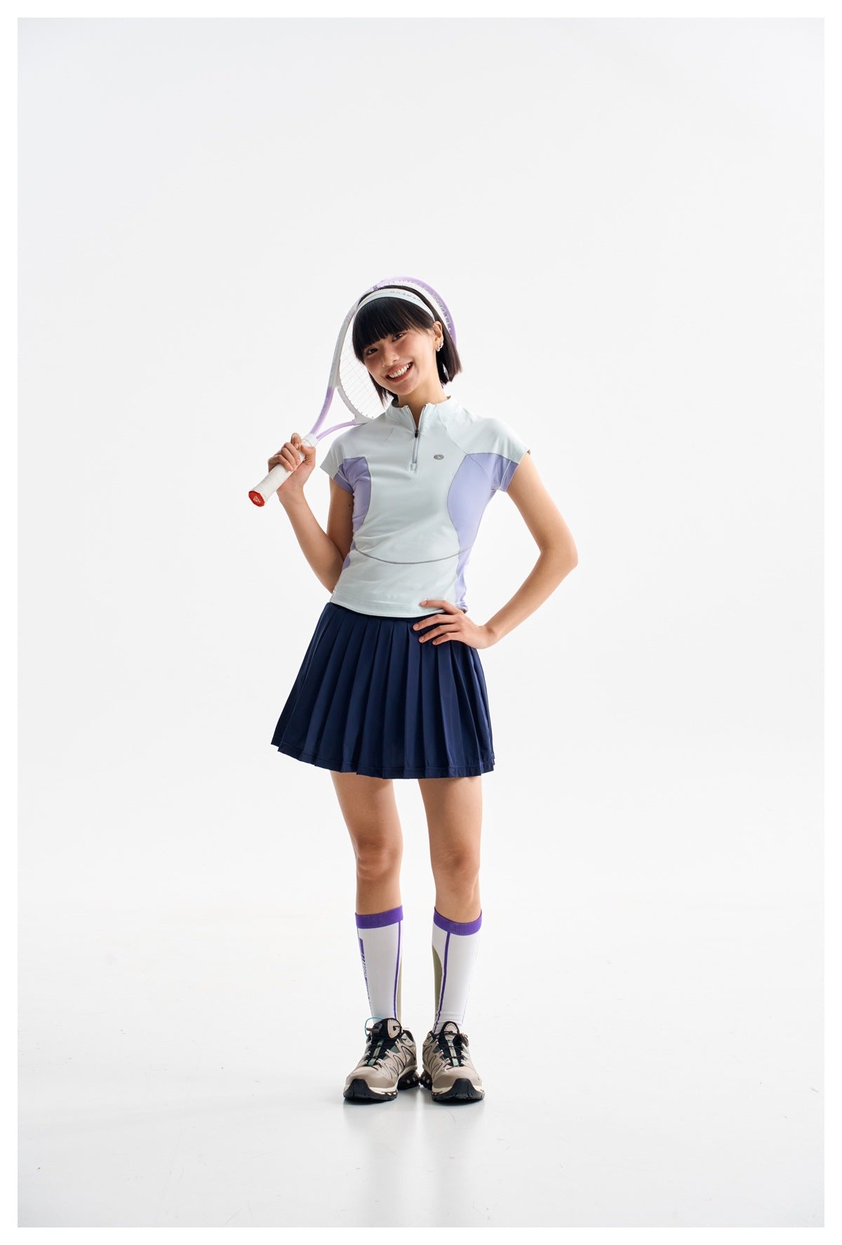 Woman in a pale blue and lavender quarter-zip top and navy pleated tennis skirt, posing with a racket over her shoulder.