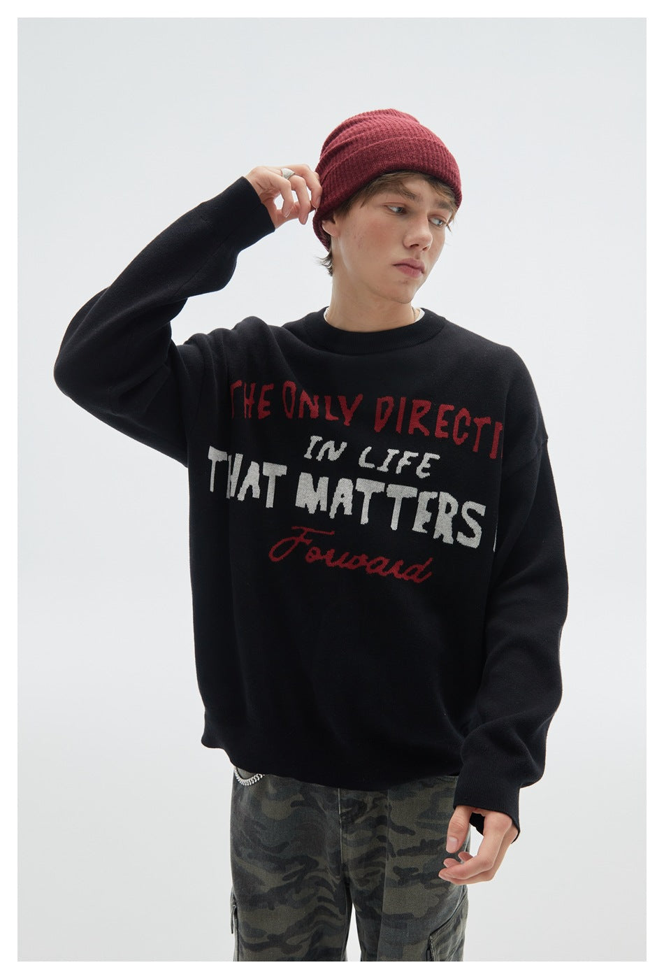 Model wearing a black crewneck sweater with red and white English lettering, a burgundy beanie, and camouflage pants.