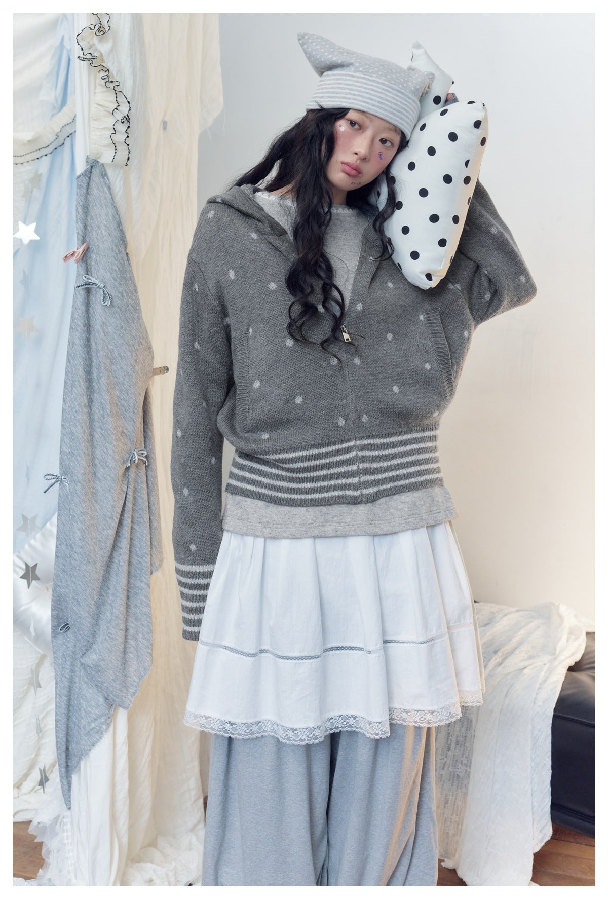 Model wears a gray patterned zip cardigan with striped ribbing over a white tiered dress, holding a polka-dot cushion.