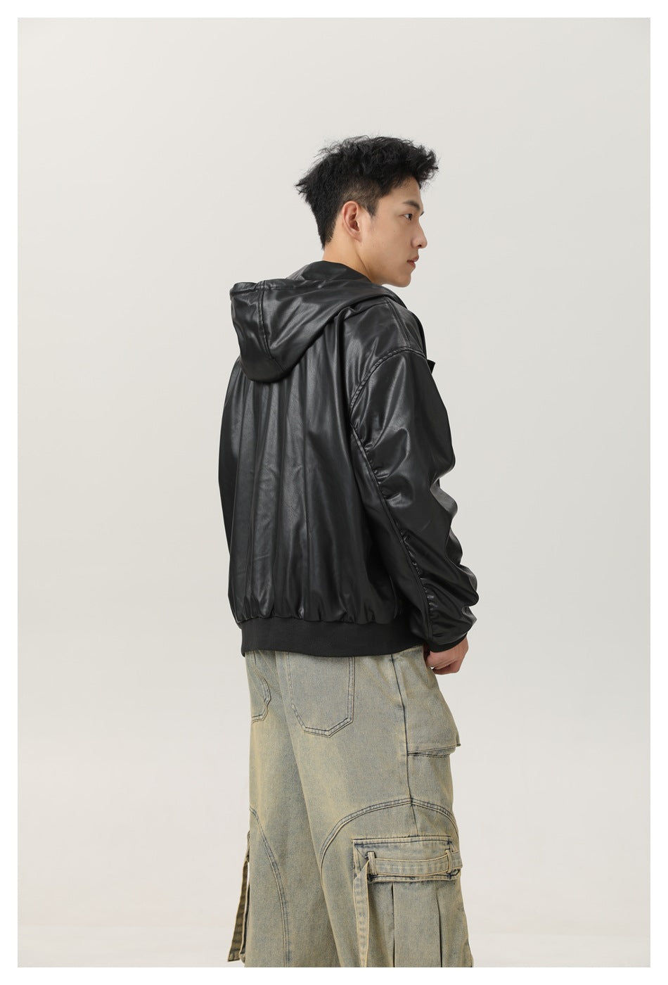 Back three-quarter view of model in black faux-leather hooded jacket with ribbed hem and wide cargo jeans