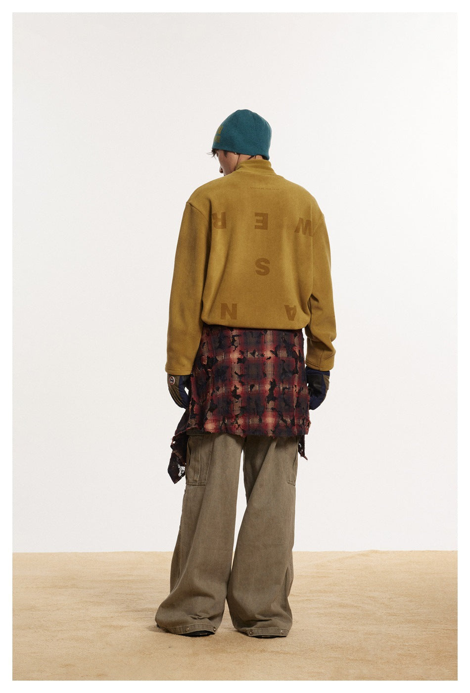 Rear view of a model wearing a mustard pullover with large scattered back lettering, teal beanie, and olive cargo pants.