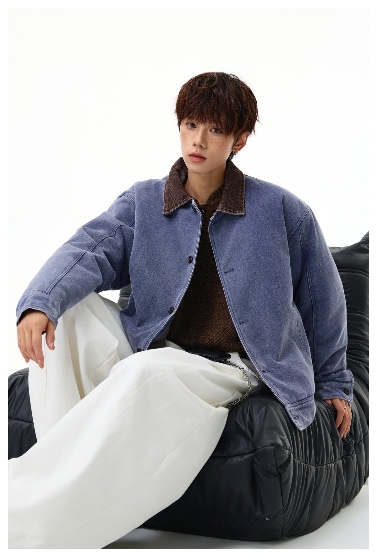 Seated model wearing a faded blue button-front jacket with a brown corduroy collar and white trousers.