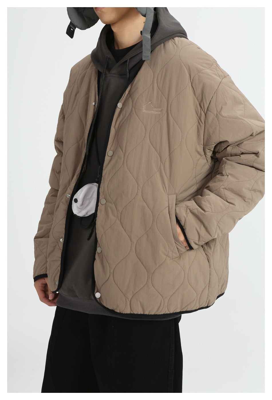Model wearing brown wavy-quilted jacket with chest embroidery, hand in pocket, over a gray hoodie