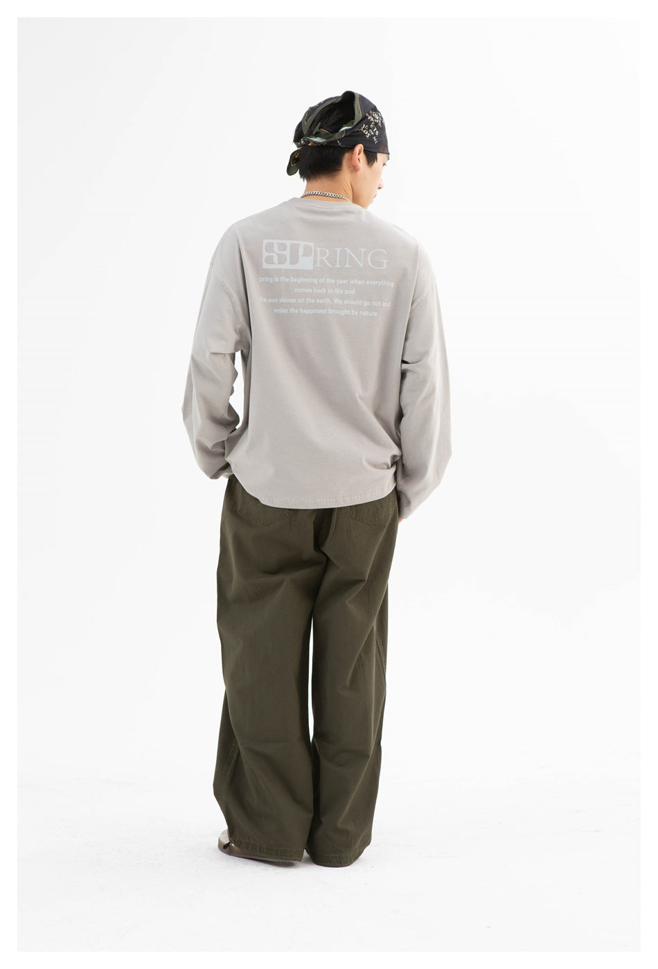 Rear view of a model wearing a gray oversized long-sleeve shirt with pale back lettering and loose olive trousers.
