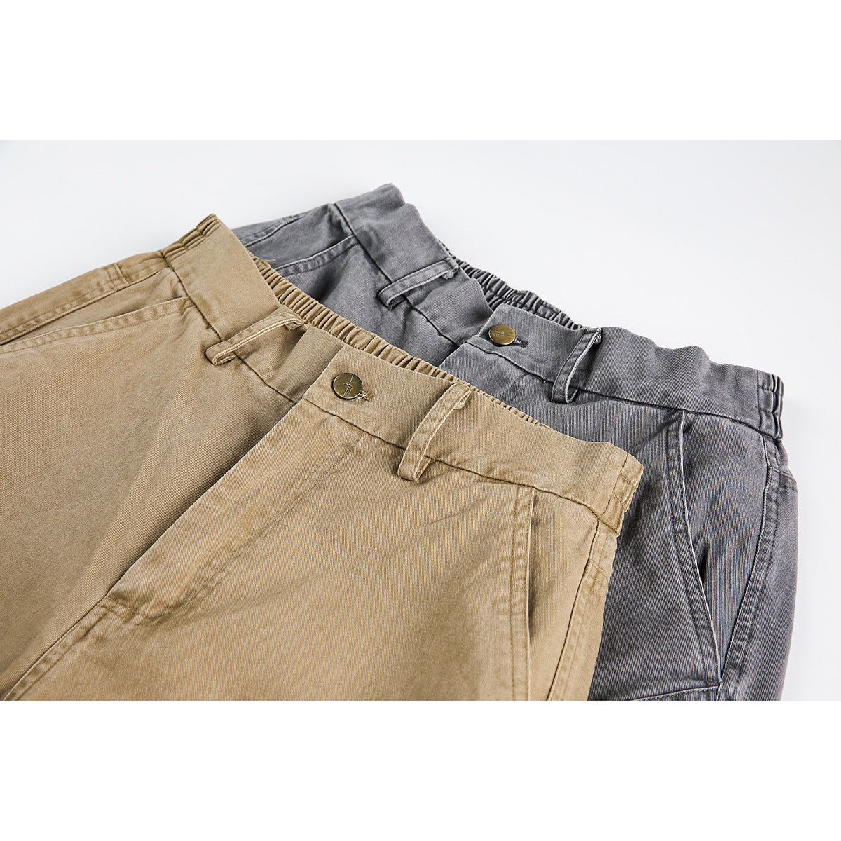 Close-up of tan and charcoal trousers overlapping, showing their front waistbands, belt loops, buttons and seams.