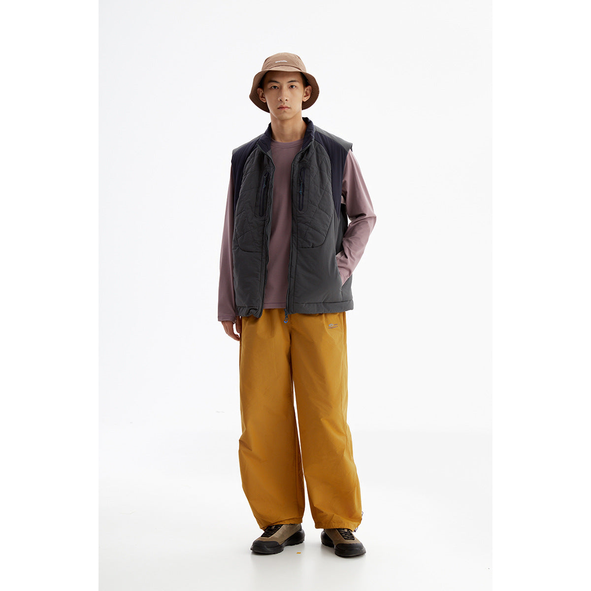 Model wearing a dark charcoal quilted vest with a mauve long-sleeve shirt and mustard pants.