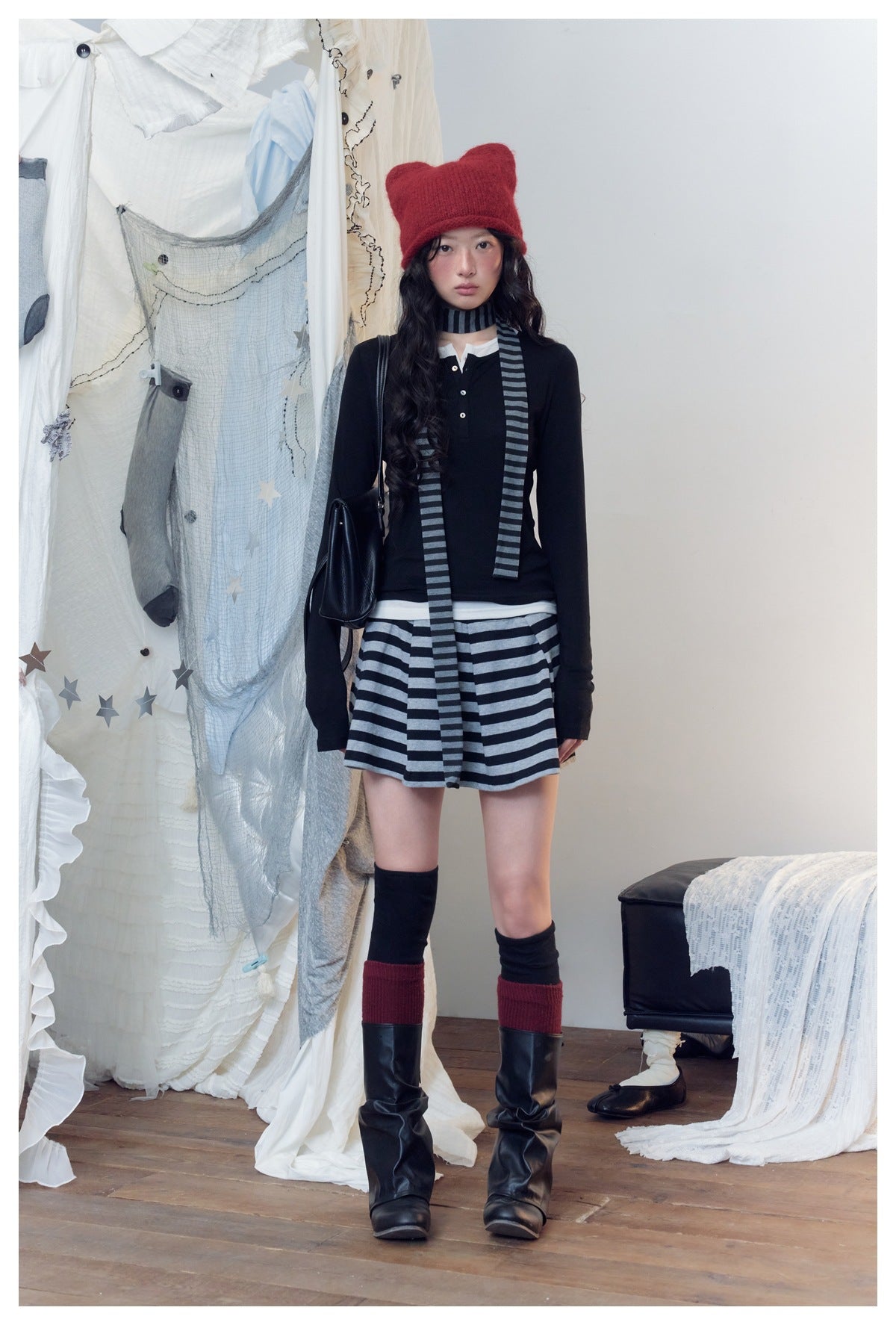 Model wears a black long-sleeve top, gray-and-black striped mini skirt and scarf, red knit hat, and tall black boots.