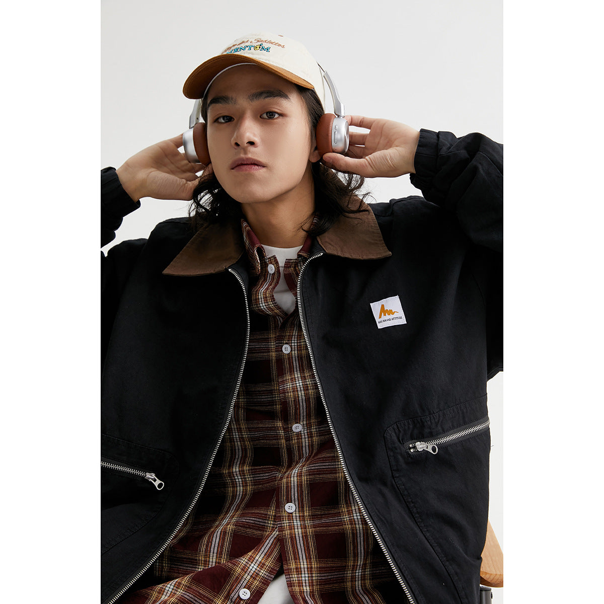 Model wearing a black zip-front jacket with a brown collar over a brown plaid shirt.