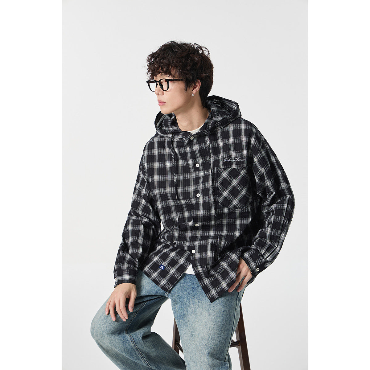 Seated model wearing a black and gray plaid hooded button-up shirt with a chest pocket.