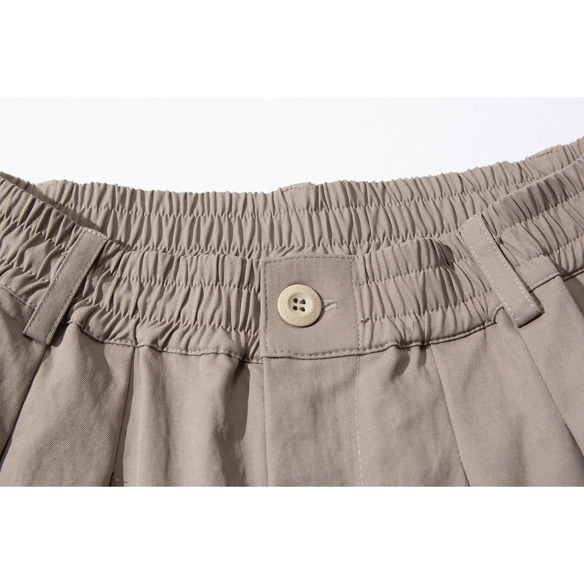Close-up of taupe shorts showing the gathered elastic waistband, belt loops, button and front pleats.