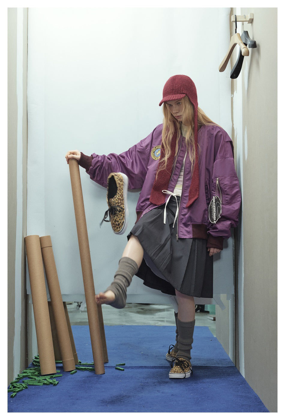 Model in a purple bomber jacket with sleeve pocket and tag, red knit hat, pleated skirt and leopard sneakers