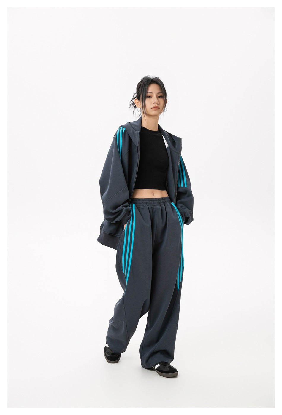 Full-length model wearing an open charcoal hoodie and loose matching pants with bright cyan stripes.