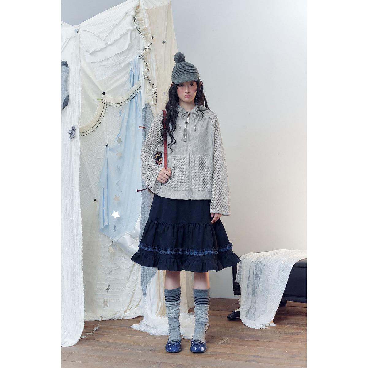 Model wearing a light gray zip hoodie with open-knit sleeves and pockets, a navy ruffle skirt, and gray leg warmers.