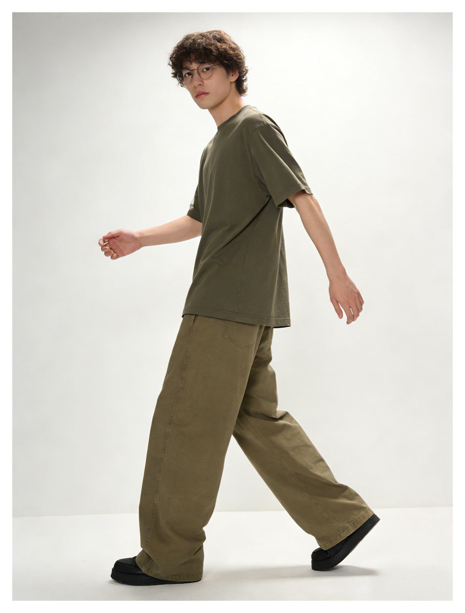 Model walking in a dark olive short-sleeve T-shirt and brown wide-leg trousers, shown from the side.