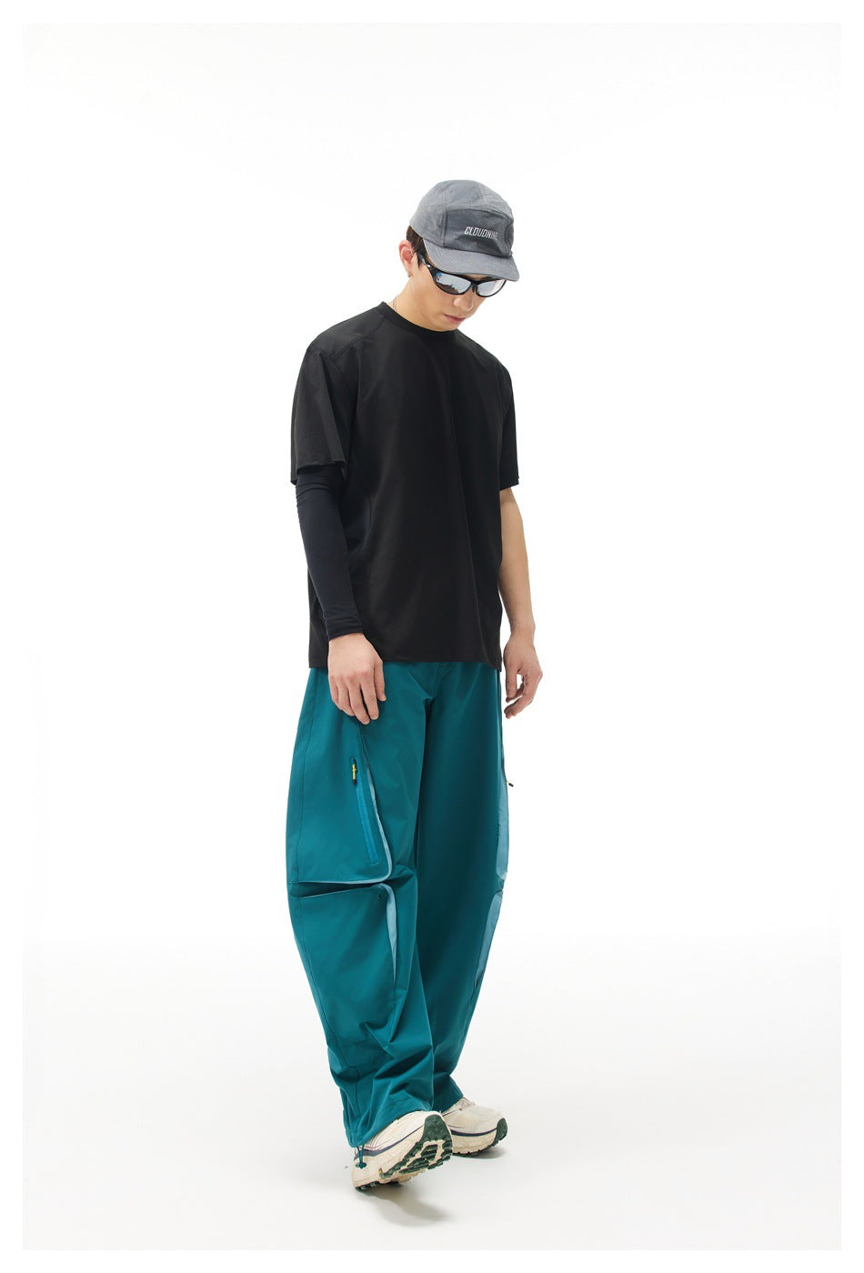 Model wearing a black short-sleeve shirt over a black sleeve and loose teal pants, shown from the front.