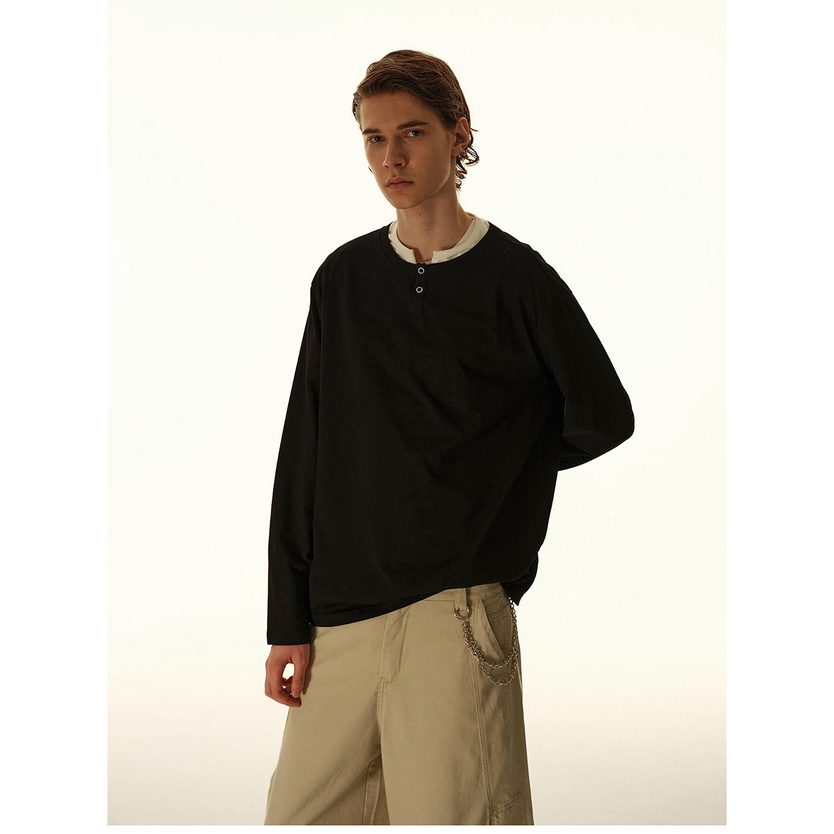 Model in a black long-sleeve henley with a white layer at the collar and loose beige trousers.