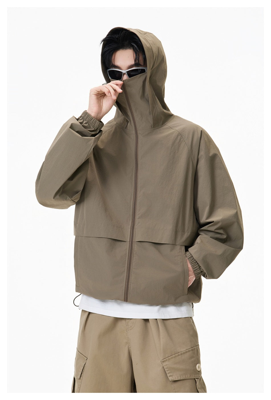 Model wearing an olive-brown hooded zip jacket with the hood up and collar raised, plus dark sunglasses.