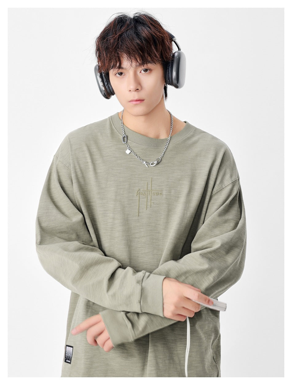 Model wearing a sage-gray oversized crewneck shirt with subtle chest embroidery and a small lower hem patch.