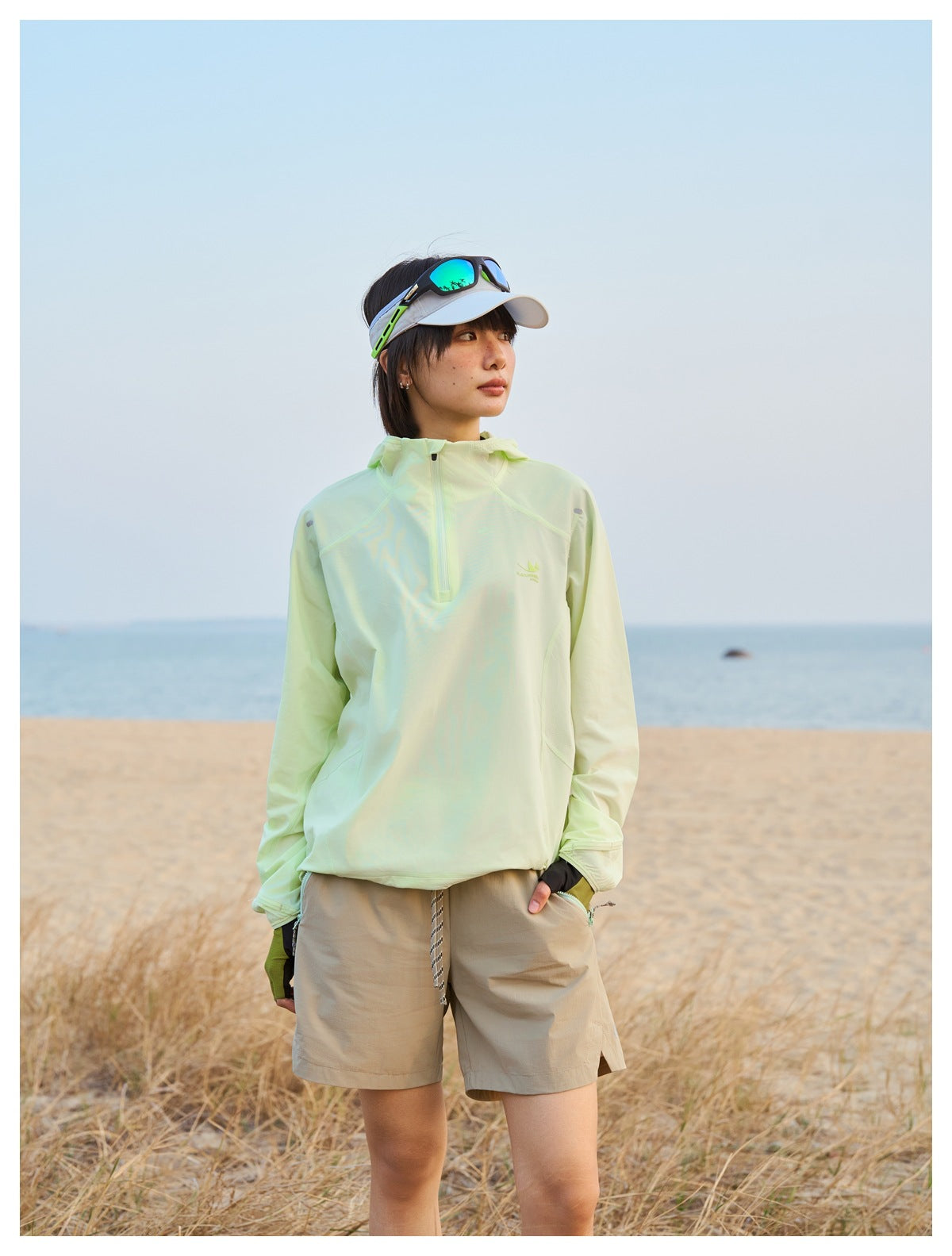 Person wearing a pale lime hooded pullover and beige shorts, standing on a beach.