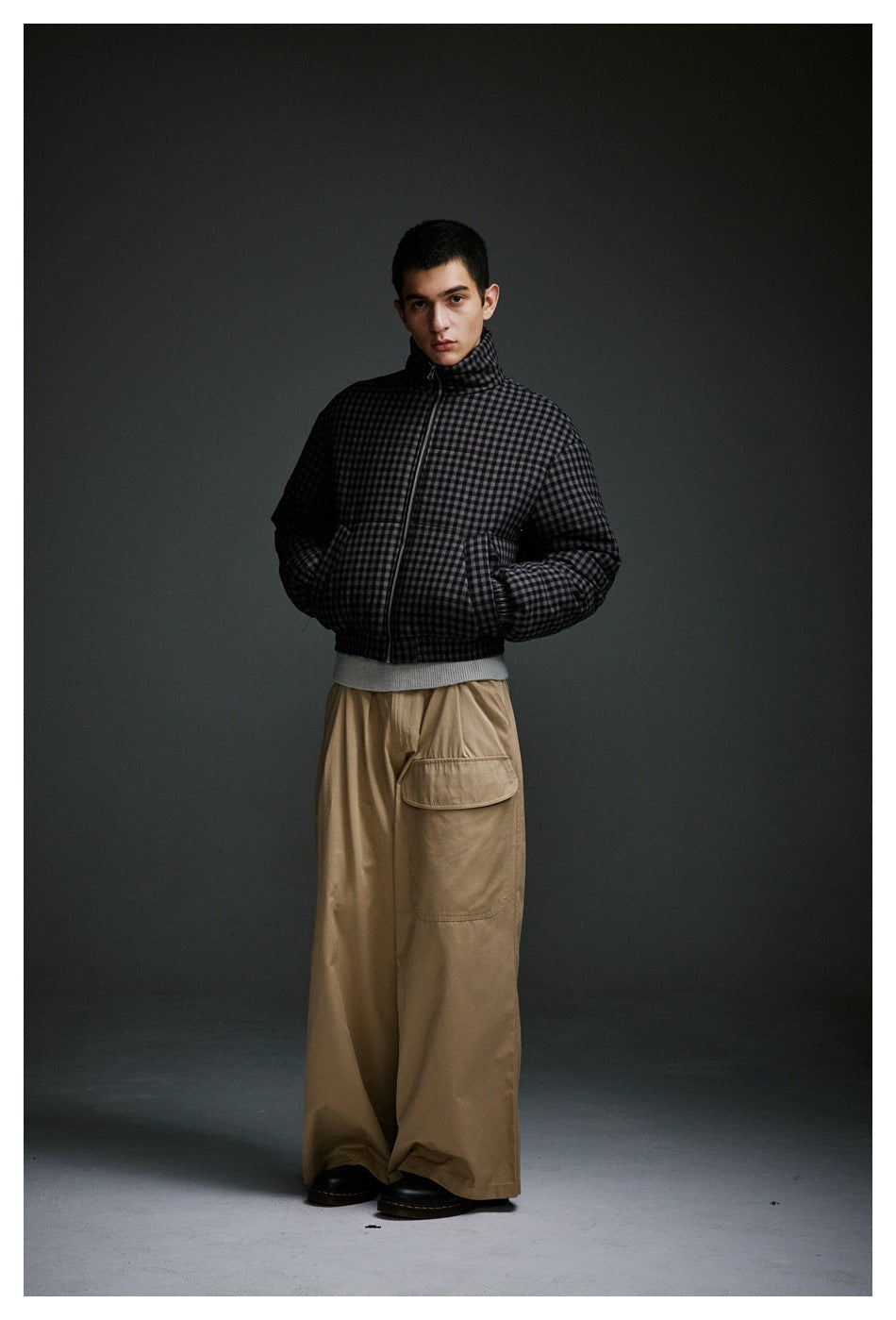 Model wearing a zipped charcoal checked padded jacket with a high collar and wide tan cargo trousers.