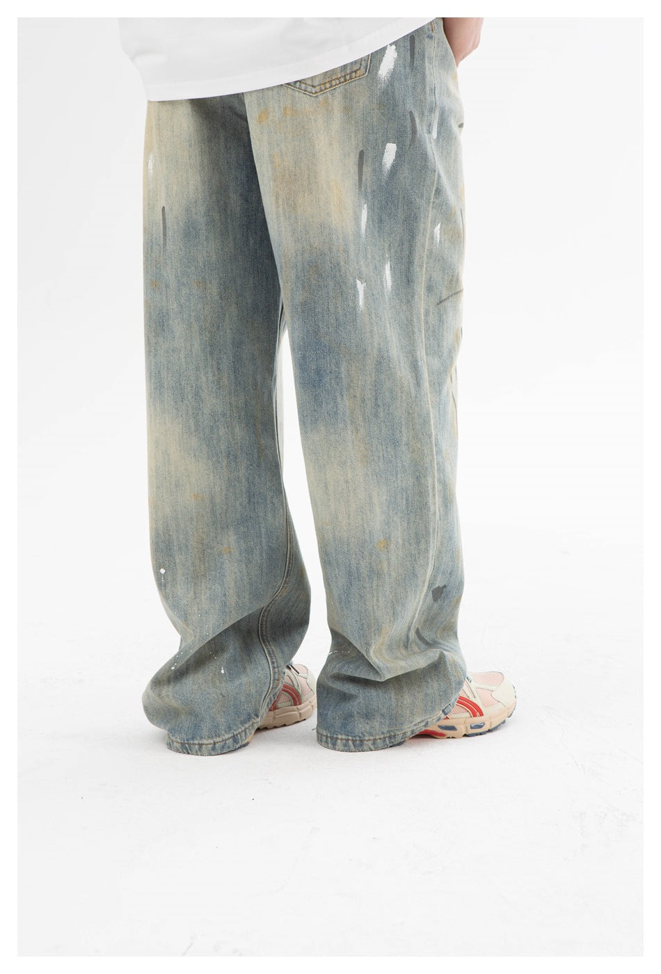 Rear view of a person wearing very loose, faded blue jeans with paint-like marks and white sneakers.