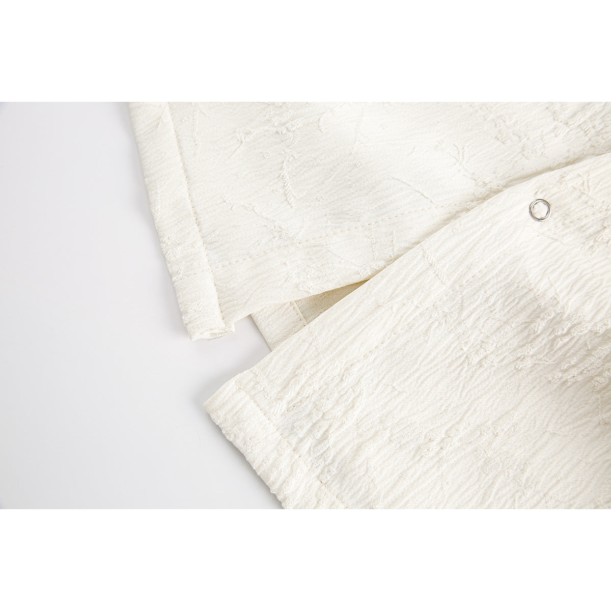 Close-up of an ivory shirt's textured fabric, sleeve hem and small circular metal fastener.
