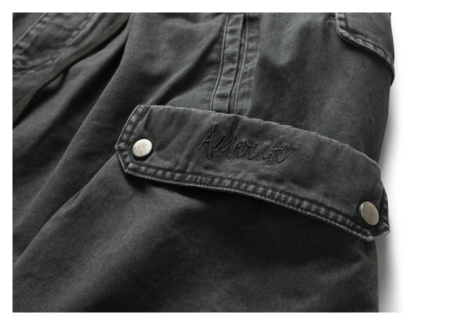 Close-up of a dark charcoal garment pocket flap with two metal snaps and script embroidery.