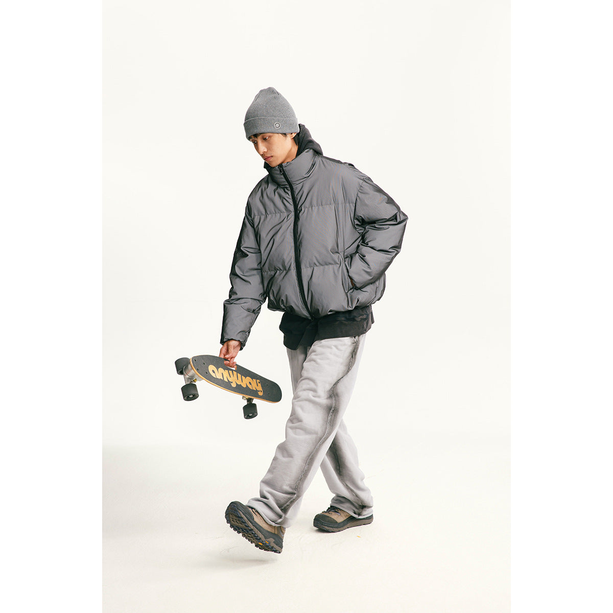 A model wears a dark gray puffer jacket and carries a skateboard in a studio.