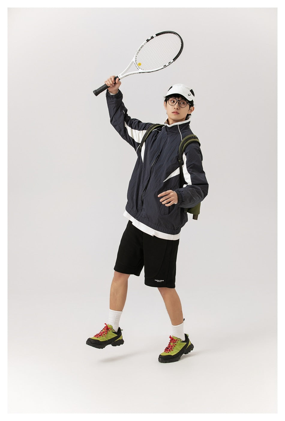 Full-length model in a dark navy jacket with white panels and black shorts, raising a tennis racket.