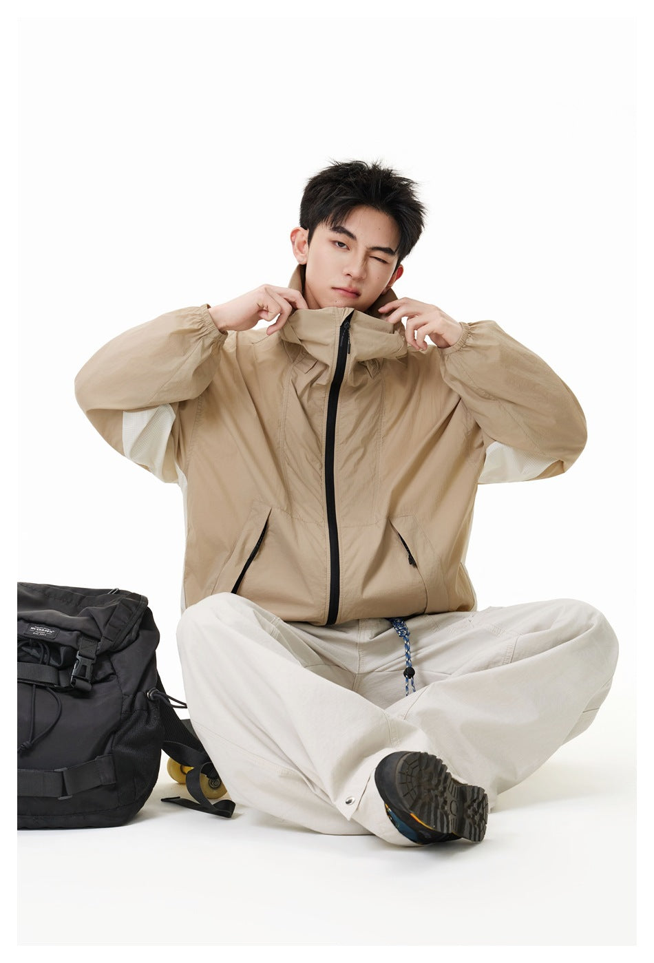 Seated model adjusting the collar of a tan high-collar zip jacket, with light trousers and a black backpack nearby.