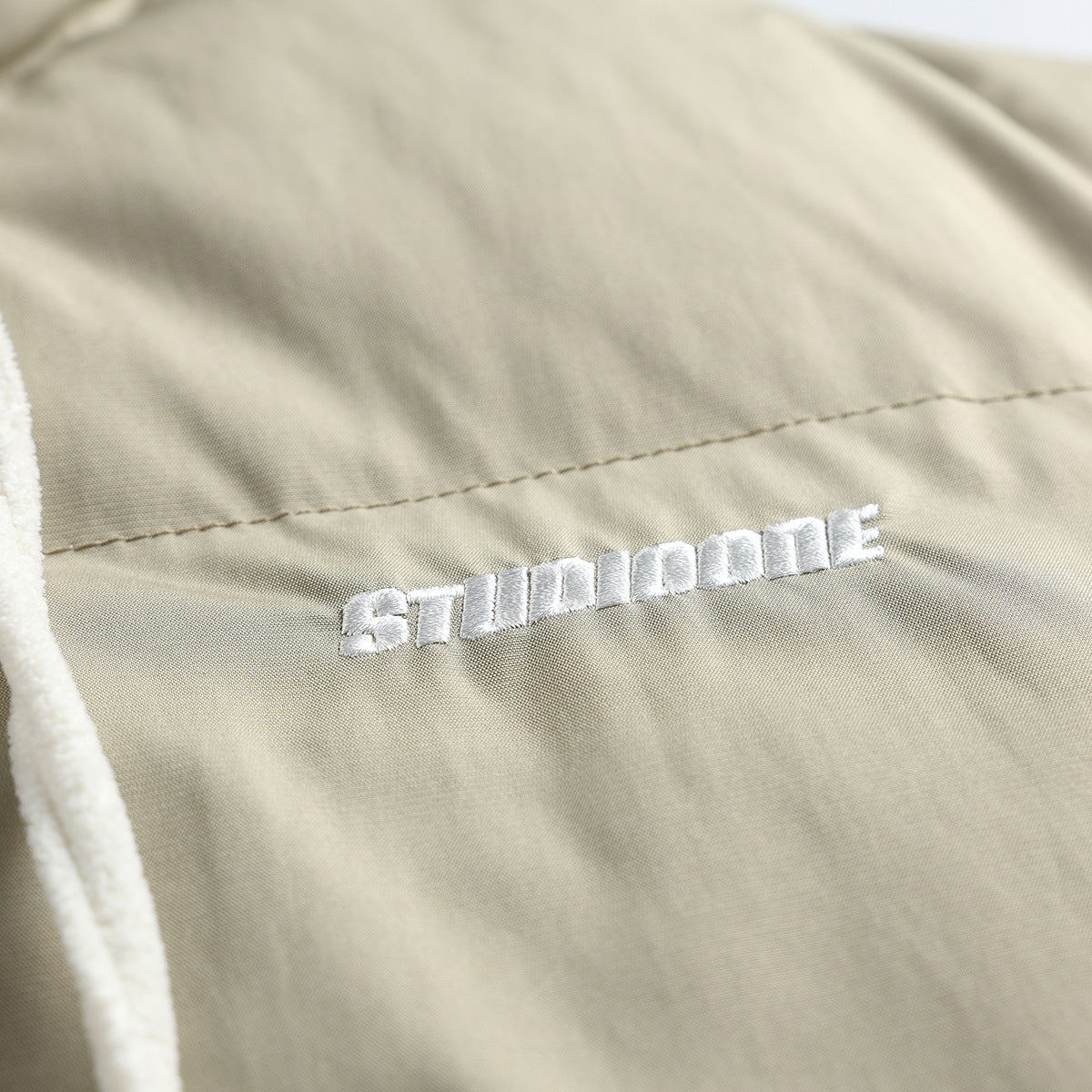 Close-up of white embroidered lettering on the taupe jacket, with a cream drawstring along the edge.
