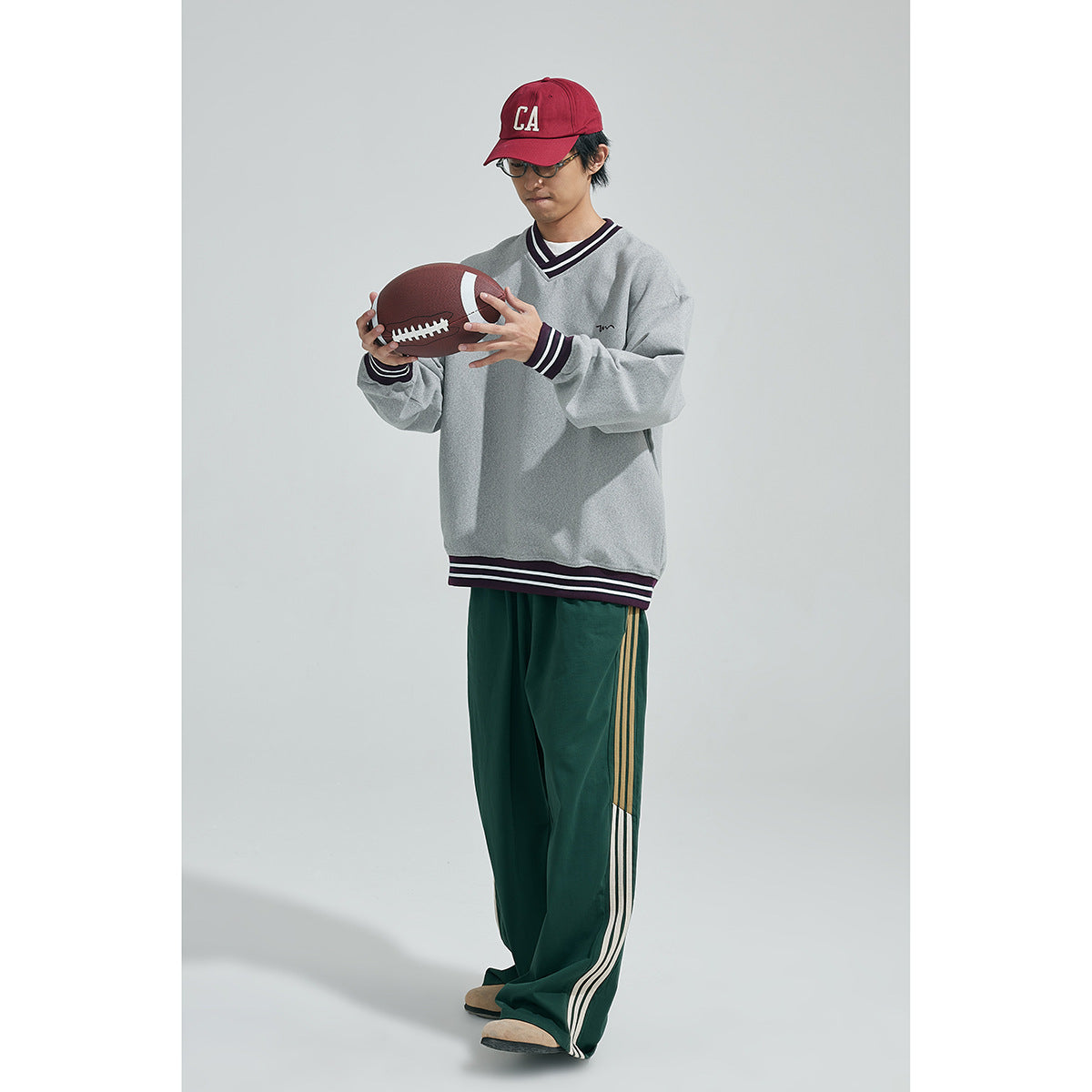 Model looking at a football while wearing a gray V-neck sweatshirt with striped trim and loose green track pants.
