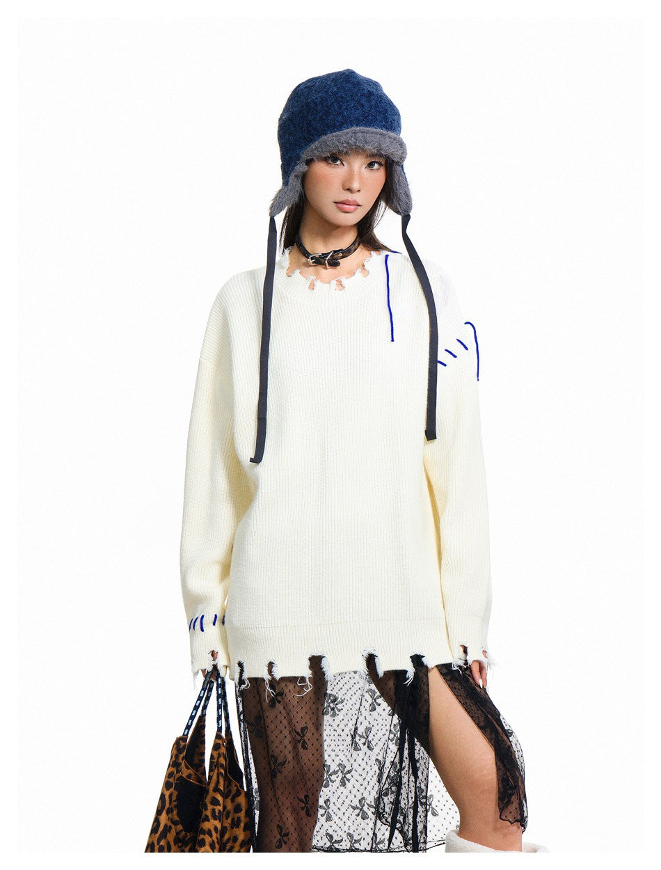 Model wearing an oversized ivory distressed sweater with blue stitch accents, a navy earflap hat, and a sheer patterned