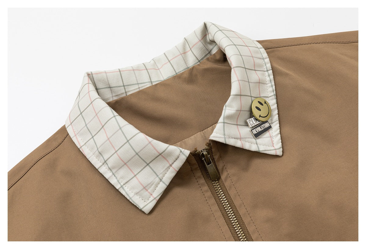 Close-up of the tan jacket's zip front, pale plaid collar, and smiley pin with text.