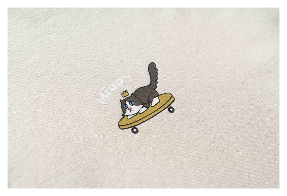 Close-up of a small cartoon cat riding a yellow skateboard with white lettering on a light beige T-shirt.