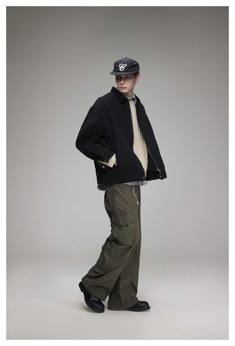 Model wearing black zip jacket with beige sweater, olive cargo pants and a cap, side pose, hand in pocket