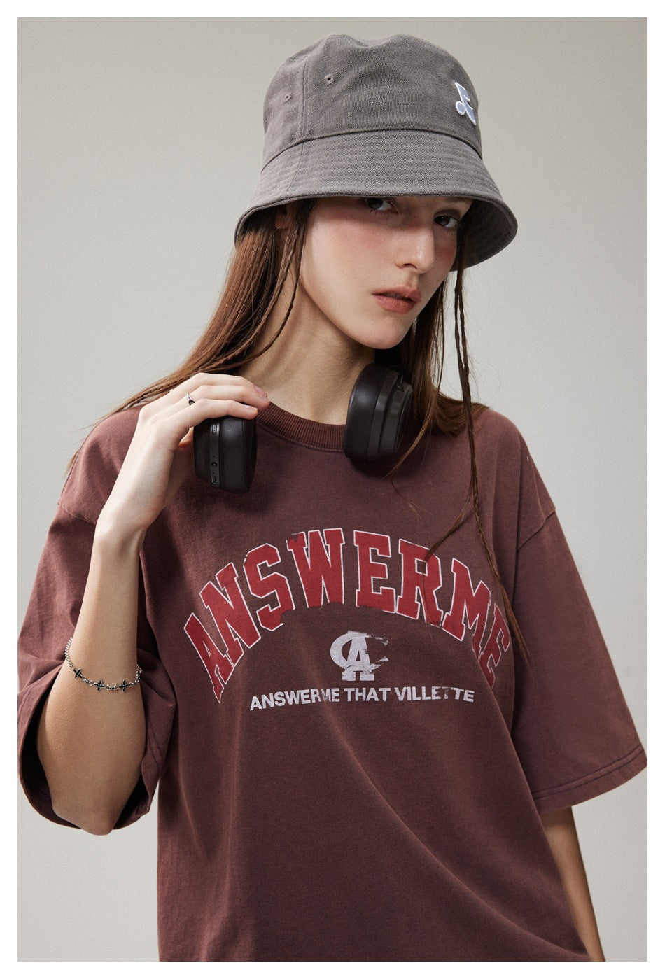 Close view of a model in a burgundy graphic T-shirt and gray bucket hat, with black headphones around her neck.