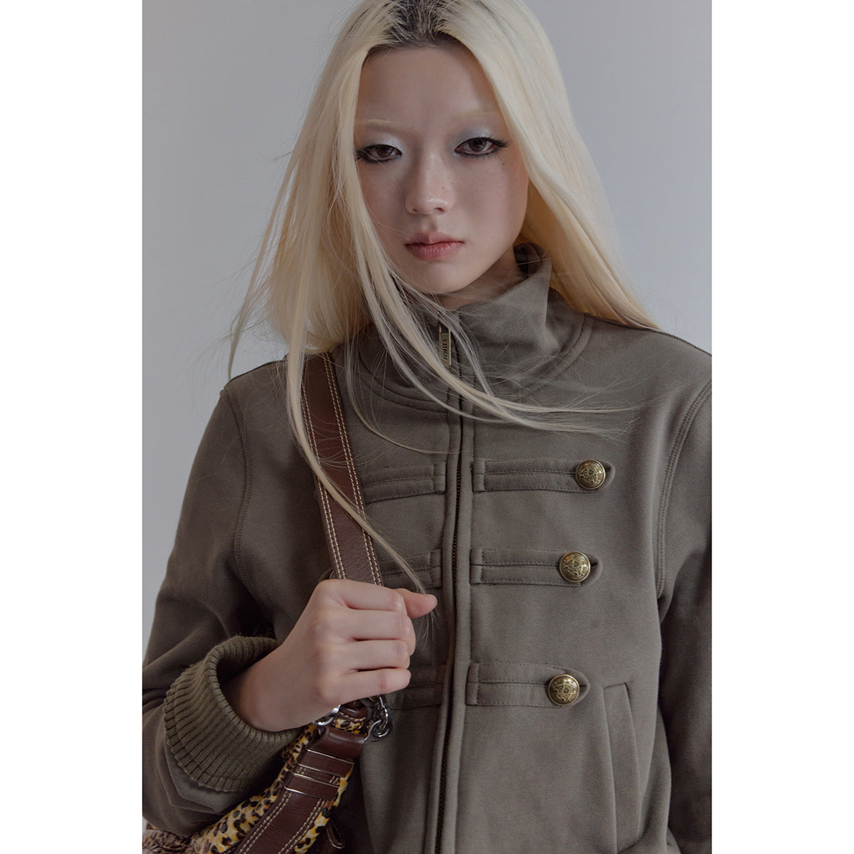 Close view of a model wearing a taupe high-collar zip jacket with brass buttons and ribbed cuffs.