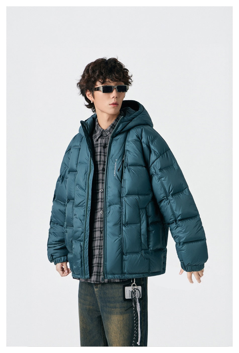 Model wearing a teal puffer jacket open over a plaid shirt with sunglasses and faded jeans, angled view