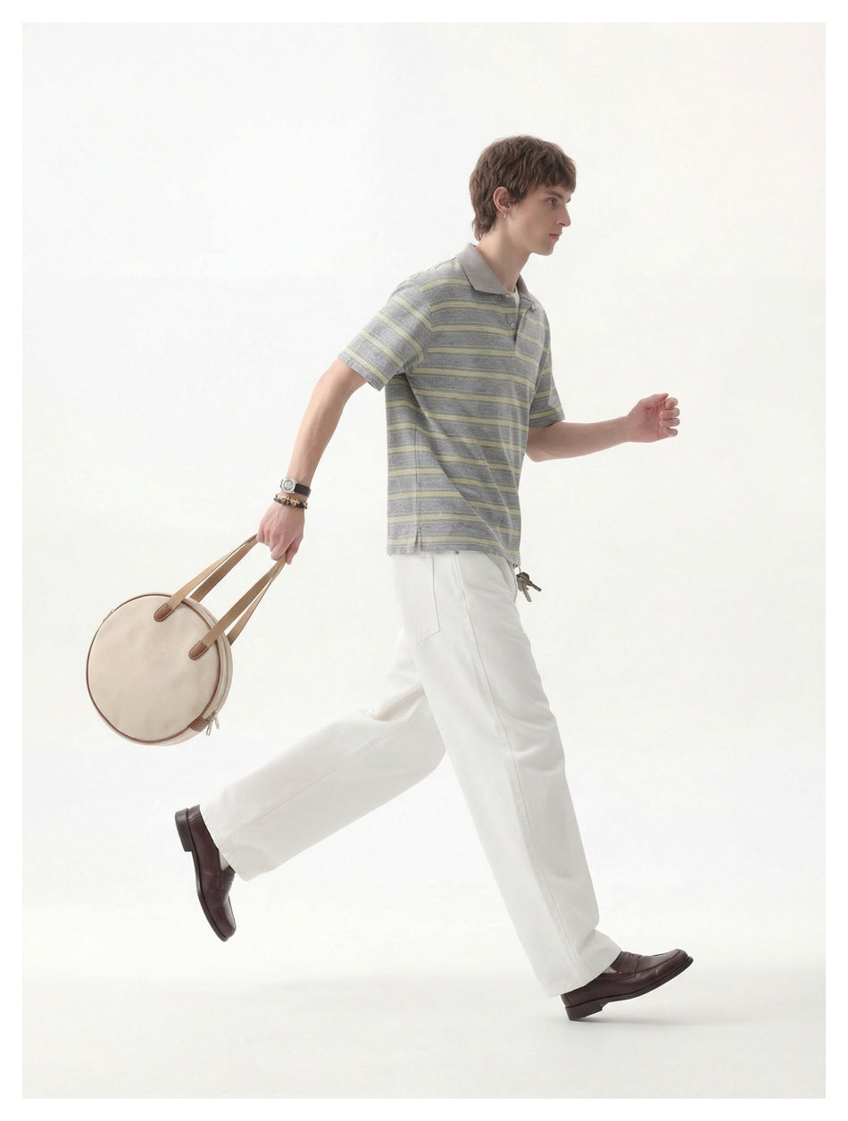 Model walking in a gray polo with pale yellow stripes and white trousers, carrying a round beige bag.