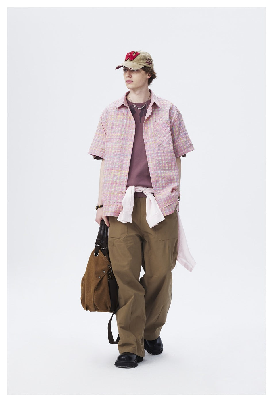 Full-length model wearing a pastel pink plaid short-sleeve shirt over a mauve tee and loose olive trousers.