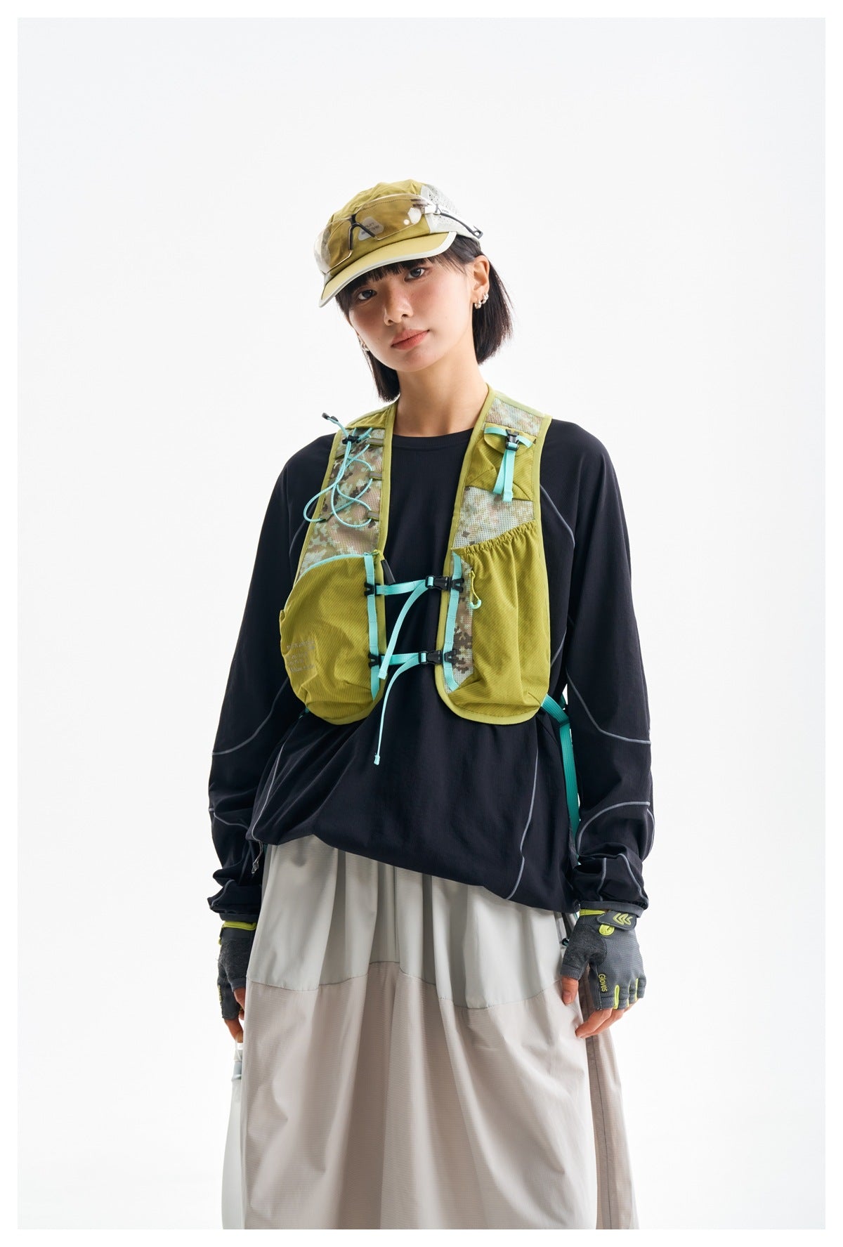 Model wearing an olive hiking vest with turquoise straps and pockets over a black top, with a cap and gloves.