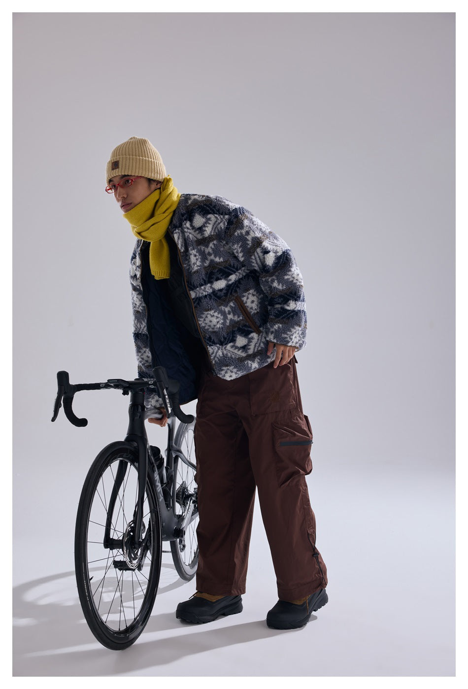 Model leans beside a bicycle in an open blue-gray and white geometric fleece jacket with brown-trimmed pockets.