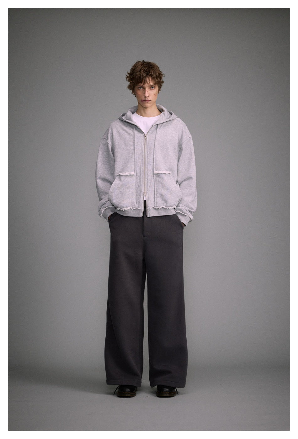 Model wearing a light gray zip-up hoodie with raw-edge pocket and hem details, paired with dark wide-leg pants.