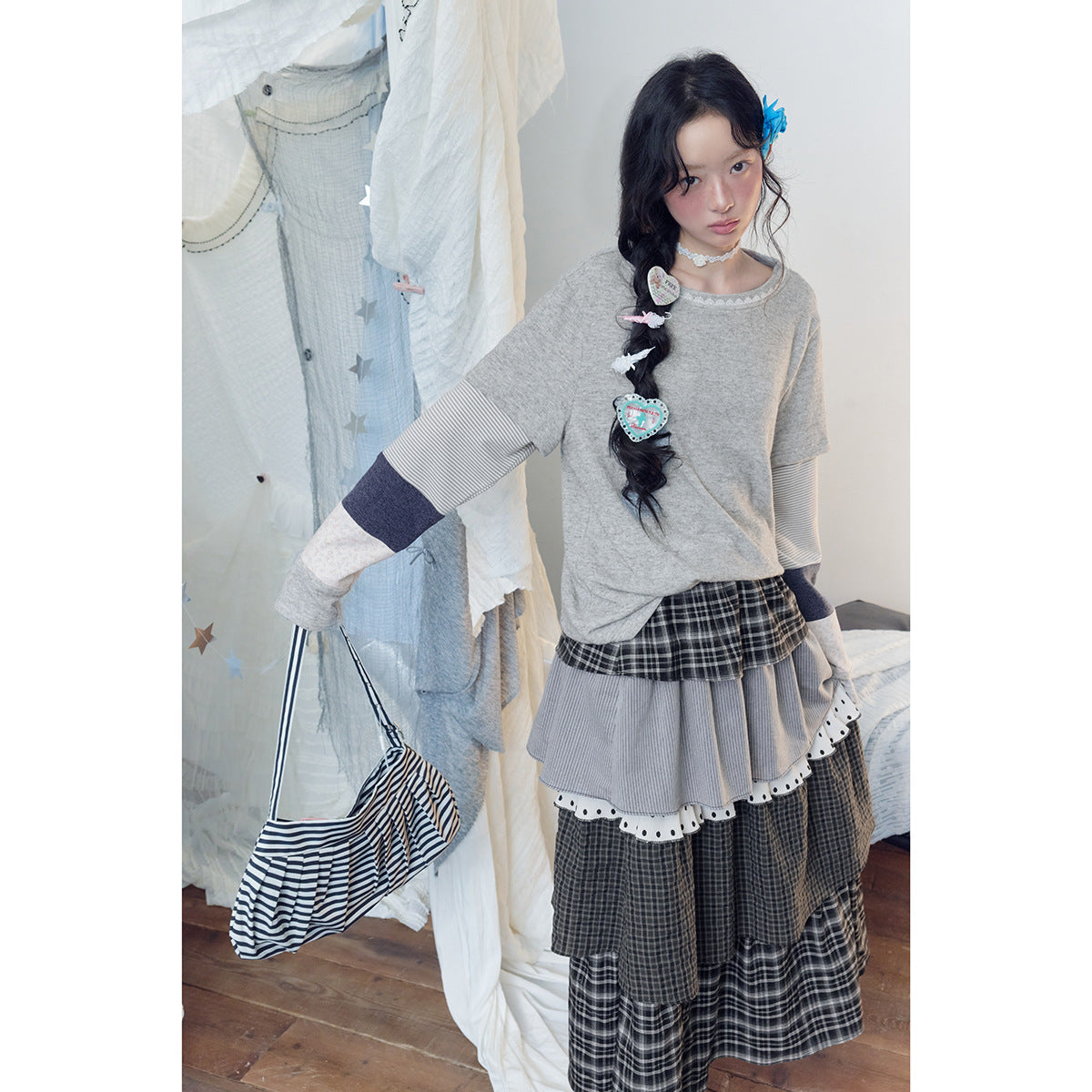 Standing model wearing a gray top with striped patchwork sleeves and a long layered plaid skirt.