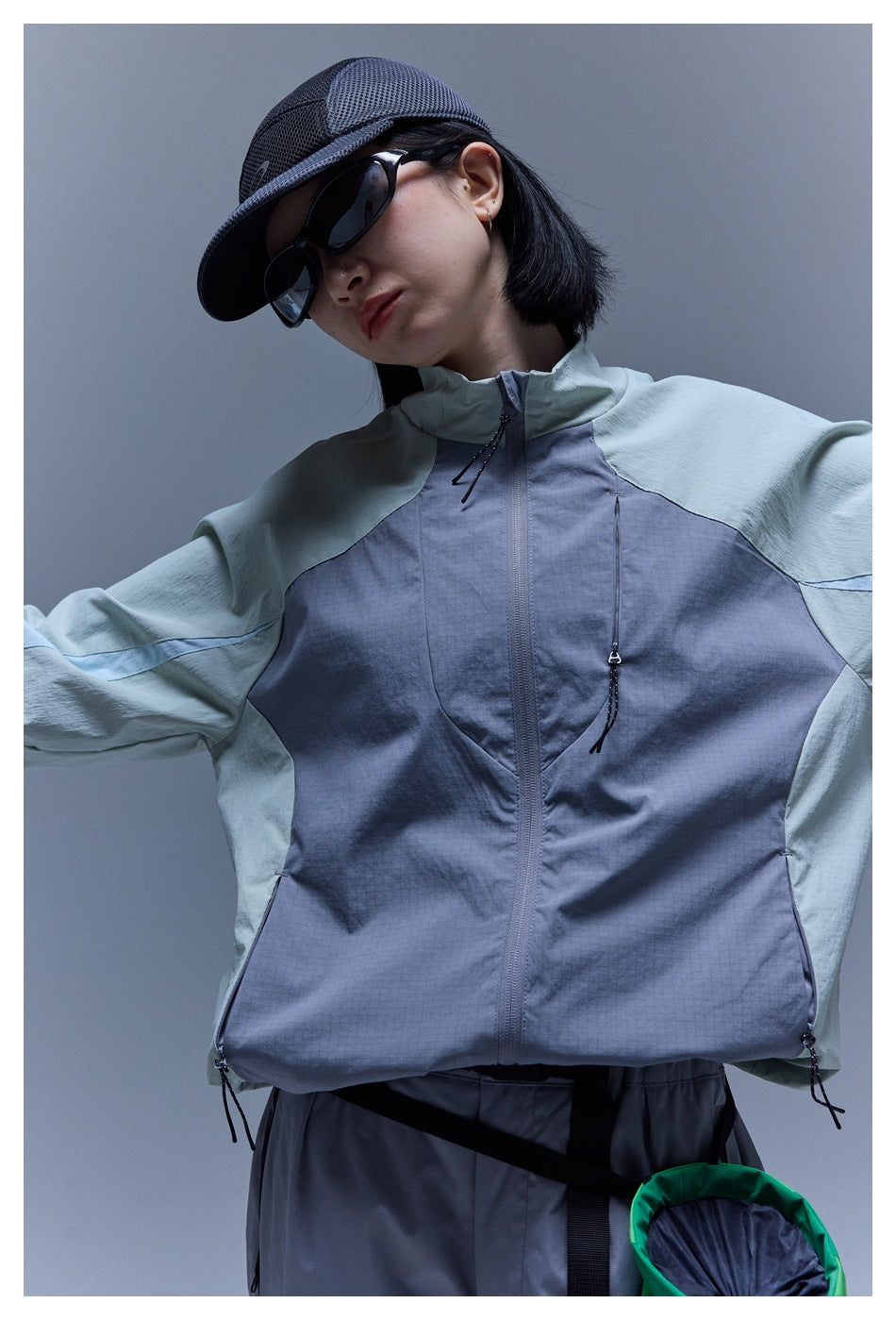 Front view of a model wearing a slate-gray and pale sage color-block jacket with a high collar and zip pockets.